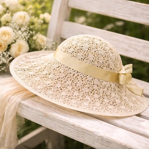 May include: A cream-colored sun hat featuring an intricate, open-weave crochet pattern throughout the crown and wide brim. A smooth, matching cream-colored satin ribbon is wrapped around the base of the crown, tied into a bow on the side.