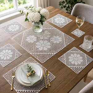 May include: A set of white crocheted lace placemats and coasters featuring a repeating floral pattern. The square placemats have a grid-like border and a central flower motif. A glass vase with white hydrangeas sits on the centre placemat.