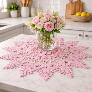 Könnte beinhalten: Rosa gehäkelte Tischdecke in Sternform mit floralem Design, zentriert auf einer Marmorarbeitsplatte. Eine klare Glasvase enthält einen Strauß rosa Rosen und anderer Blumen. Die Küchenszene umfasst weiße Schränke und einen Korb mit Zitronen.