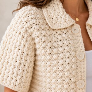 Puede incluir: Un c&aacute;rdigan de ganchillo color crema con un patr&oacute;n de textura de punto bobble, mangas cortas, cuello cl&aacute;sico y tres botones grandes, redondos y a juego en la parte delantera. La prenda est&aacute; confeccionada con un hilo denso y uniforme.