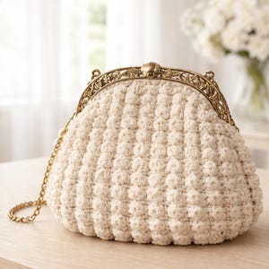 May include: Cream-colored, textured clutch with a gold-toned metal frame and chain strap. The purse features a rounded shape and a detailed clasp. The fabric has a raised, bobble-like texture. A stylish accessory for any occasion.