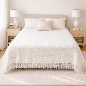 May include: A white bedspread with a geometric pattern and fringe trim. The bed is made up with white pillows and a decorative pillow. Two white lamps sit on matching nightstands on either side of the bed.
