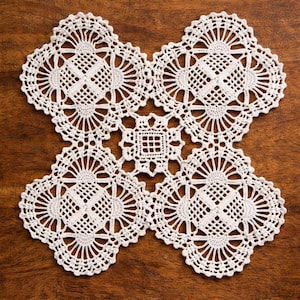 Puede incluir: Un tapete blanco hecho a mano con ganchillo con un diseño simétrico. El tapete presenta un cuadrado central rodeado de cuatro secciones más grandes, de forma cuadrada, cada una con un patrón calado detallado. El tapete está colocado sobre una superficie de madera.