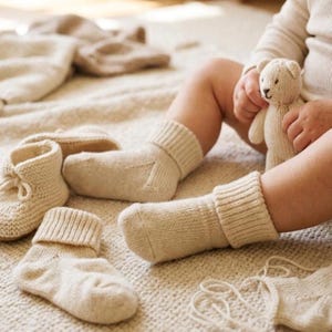 May include: A set of cream-colored knitted baby items including a pair of cuffed socks, a pair of booties with tie closures, and a small knitted teddy bear. The items feature a simple stockinette stitch pattern and ribbed cuffs on the socks.
