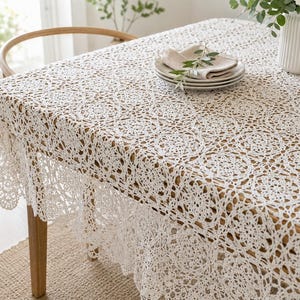 May include: A white rectangular tablecloth featuring an intricate, repeating circular crochet pattern with scalloped edges. The lace fabric drapes over the sides of a wooden table, which is topped with a stack of plates and a white ceramic vase.