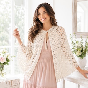 May include: A cream-colored, open-front crochet capelet featuring an intricate, airy diamond-patterned knit. The garment has a round neckline with a simple tie closure made of matching yarn, finished with small tassels at the ends of the ties.