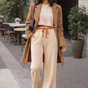 May include: A camel-coloured wool coat worn over a beige knit crop top and matching wide-leg knit trousers with an orange drawstring waist. The outfit includes white trainers, a dark brown shoulder bag, and dark-tinted sunglasses.