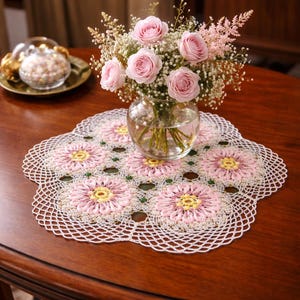 May include: A round white crocheted doily featuring a floral pattern with pink and yellow centers, displayed under a clear glass vase filled with a bouquet of pink roses and small white filler flowers, creating a decorative centerpiece.