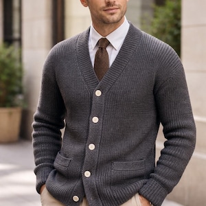 Pode incluir: Um cardigan cinza carvão com decote em V, fecho de botões e dois bolsos frontais. O suéter é usado sobre uma camisa branca e uma gravata marrom. A pessoa também está usando calças cáqui.