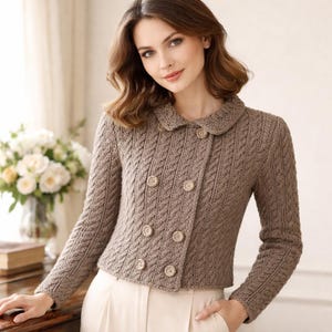 May include: A light brown, double-breasted knit cardigan featuring a cable knit pattern, a rounded collar, and six matching buttons. The garment has long sleeves and a cropped length, providing a structured look with a textured, woven finish.