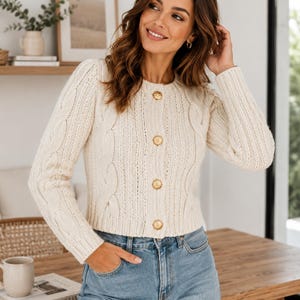 Könnte beinhalten: Eine cremefarbene Strickjacke mit Zopfmuster, langen Ärmeln, Rundhalsausschnitt und vier goldfarbenen, runden Knöpfen auf der Vorderseite. Der Pullover wird mit einer hellblauen Jeans kombiniert und zeigt eine strukturierte Optik mit vertikalen Zopfmustern.