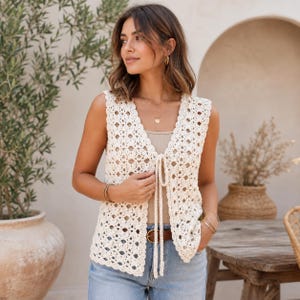 May include: A sleeveless, off-white crochet vest featuring an intricate diamond-shaped openwork pattern. The garment has a V-neckline and is secured at the front with two long, matching crochet ties that hang down from the center of the bodice.