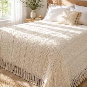 Op de afbeelding: Crèmekleurig sprei met een geometrisch patroon en franjes. De sprei is gedrapeerd over een bed met witte kussens en een decoratief kussen. Een vaas met groen staat op een houten nachtkastje.