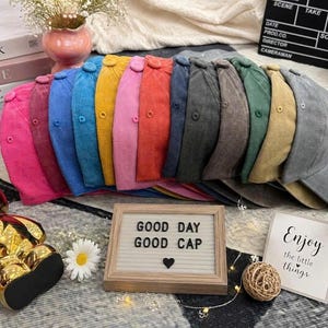 May include: A collection of twelve baseball caps in a variety of colors including pink, blue, yellow, red, and gray, arranged in a row. Each cap features a button on top and eyelets. A wooden letter board displays the text GOOD DAY GOOD CAP.