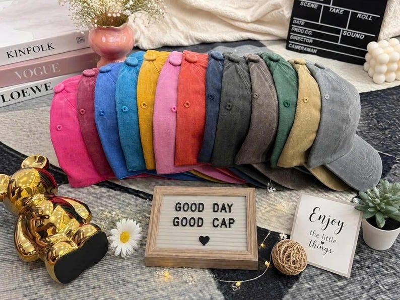May include: A collection of twelve baseball caps in a variety of colors including pink, blue, yellow, red, and gray, arranged in a row. Each cap features a button on top and eyelets. A wooden letter board displays the text GOOD DAY GOOD CAP.