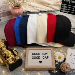May include: A collection of seven baseball caps in burgundy, blue, navy, white, cream, red, and black, arranged in a row. A rectangular letter board in front displays the text GOOD DAY GOOD CAP with a heart symbol, alongside a gold bear figurine.