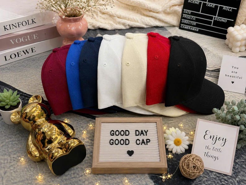 May include: A collection of seven baseball caps in burgundy, blue, navy, white, cream, red, and black, arranged in a row. A rectangular letter board in front displays the text GOOD DAY GOOD CAP with a heart symbol, alongside a gold bear figurine.