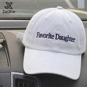 May include: A white baseball cap featuring the text Favorite Daughter embroidered in dark blue thread across the front panel. The cap has a structured crown, a curved brim with visible stitching, and a fabric strap closure at the back.