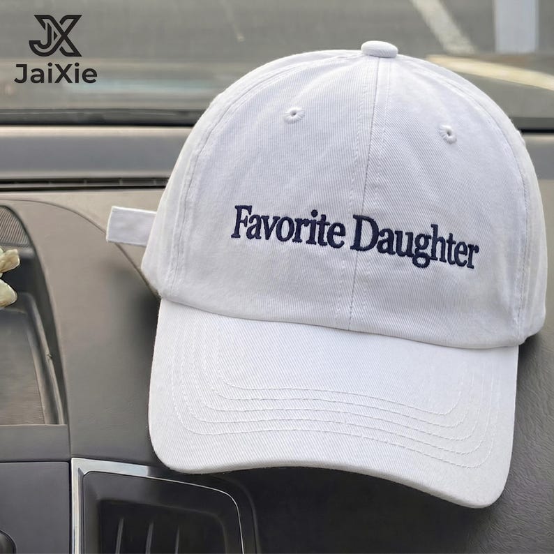 May include: A white baseball cap featuring the text Favorite Daughter embroidered in dark blue thread across the front panel. The cap has a structured crown, a curved brim with visible stitching, and a fabric strap closure at the back.