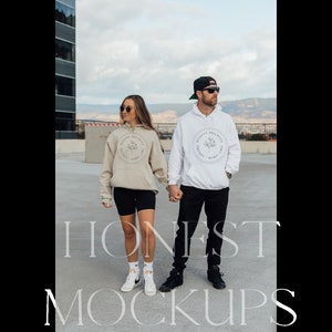 May include: A couple wearing matching hoodies with a floral design and the text "Honest" in a circle. The woman is wearing a beige hoodie and black shorts, while the man is wearing a white hoodie and black trousers. They are standing on a rooftop with a city skyline in the background.