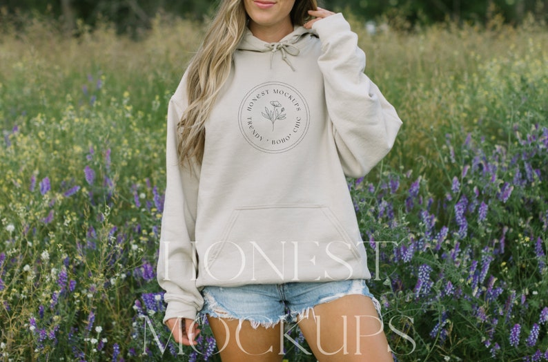 May include: A beige hoodie with a front pocket and a drawstring hood. The hoodie features a circular logo with text and a floral design. The person is wearing denim shorts. The background is a field of purple and green wildflowers.