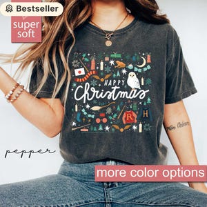 May include: Dark grey t-shirt featuring a "Happy Christmas" design with festive illustrations. The design includes an owl, a broom, and other holiday-themed elements. The shirt has a "super soft" label.