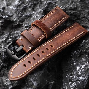 May include: A brown leather watch strap with white stitching. The strap has a buckle and is designed for a wristwatch.