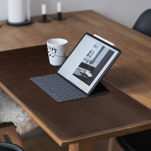 May include: A brown leather desk mat with a stitched edge, a white wireless keyboard, and a tablet computer with a white stylus on a wooden table.