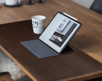 May include: A brown leather desk mat with a stitched edge, a white wireless keyboard, and a tablet computer with a white stylus on a wooden table.