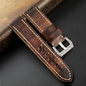 May include: A brown leather watch strap with white stitching and a silver buckle. The buckle has a decorative design.