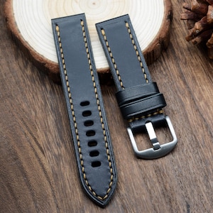 May include: Two-piece black leather watch strap with contrasting yellow stitching. The strap features a stainless steel buckle and multiple adjustment holes. The watch band is displayed on a wooden surface.