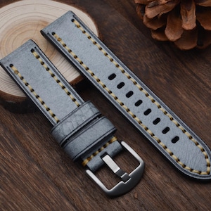 May include: A pair of gray leather watch straps with yellow stitching. The straps have a silver buckle and are displayed on a wooden surface. The watch straps are a stylish accessory.