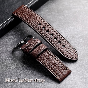 Op de afbeelding: Een bruine bison leren horlogeband met een getextureerd oppervlak en witte stiksels. De band heeft meerdere gaten voor aanpassing en een zilverkleurige gesp. De tekst "Bison leather strap" is zichtbaar.