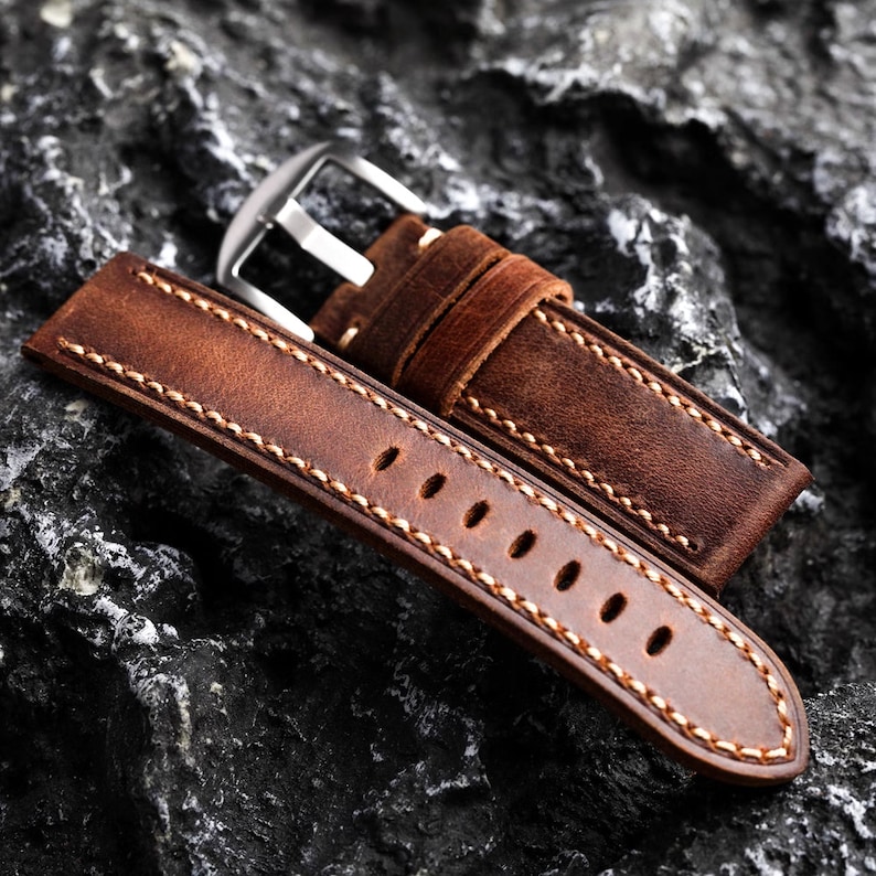 May include: A brown leather watch strap with white stitching and a silver buckle.