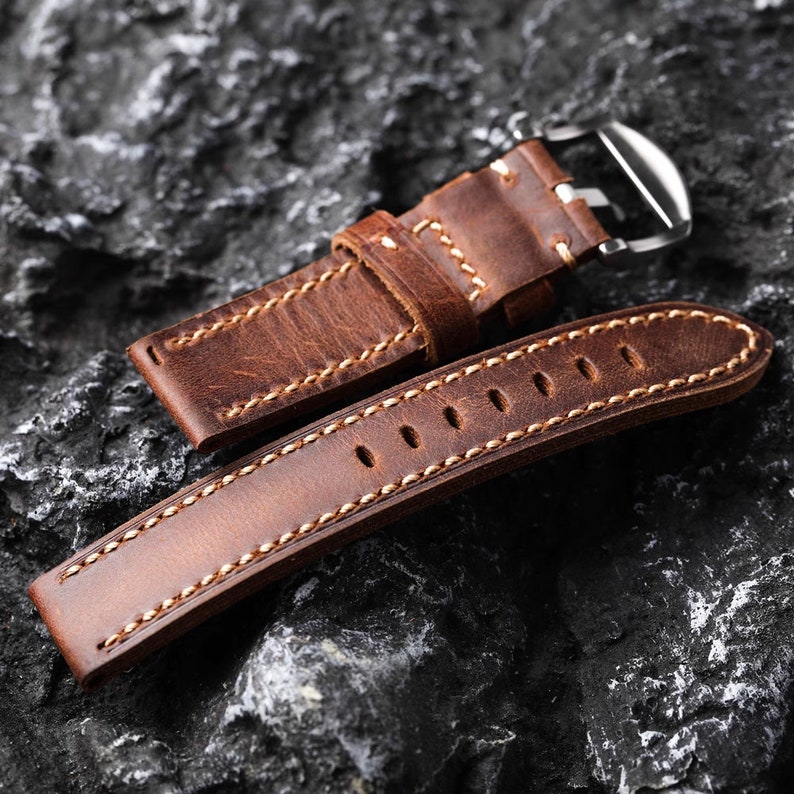 May include: A brown leather watch strap with white stitching and a silver buckle. The strap is laid out on a dark gray textured surface.