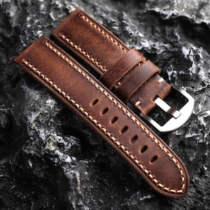 May include: A brown leather watch strap with white stitching and a silver buckle. The strap is lying on a dark gray textured surface.