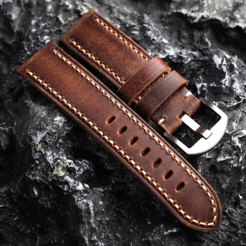May include: A brown leather watch strap with white stitching and a silver buckle. The strap is lying on a dark gray textured surface.