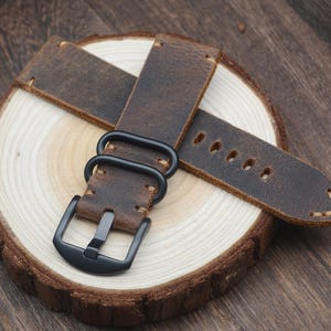May include: A brown leather watch strap with a black buckle and hardware. The strap is distressed, showing wear and a rugged texture. The watch band is displayed on a wooden slice, showcasing the natural grain and color variations.