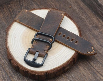 May include: A brown leather watch strap with a black buckle and hardware. The strap is distressed, showing wear and a rugged texture. The watch band is displayed on a wooden slice, showcasing the natural grain and color variations.