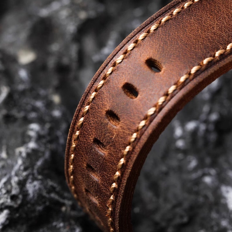 May include: Close-up of a brown leather watch strap with a light brown stitching detail. The strap has multiple holes for adjusting the fit.