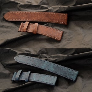 Op de afbeelding: Twee leren horlogebandjes. E&eacute;n is bruin met een verweerde afwerking en zichtbare stiksels. De andere is blauw met een verweerde afwerking en zichtbare stiksels. Beide bandjes hebben een gesp en worden gepresenteerd op een donkere, getextureerde ondergrond.