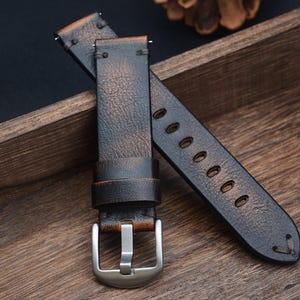 May include: A distressed brown leather watch strap with a silver buckle. The strap has a vintage look with visible wear and tear. The strap is approximately 9 inches long.