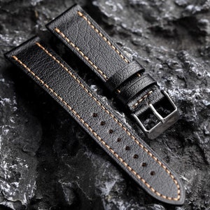 May include: Two black leather watch straps with orange stitching. The straps are laid out on a textured gray surface.