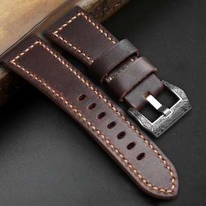 Puede incluir: Primer plano de una correa de reloj de cuero marrón con costuras color crema. La correa tiene una hebilla plateada con un diseño ornamentado. El cuero parece de alta calidad y duradero, para un reloj.