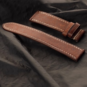 Op de afbeelding: Twee bruine leren horlogebandjes met witte stiksels. De bandjes zijn gemaakt van een distressed leermateriaal en zijn ontworpen om aan een horloge te worden bevestigd. De bandjes worden op een donkere achtergrond getoond.