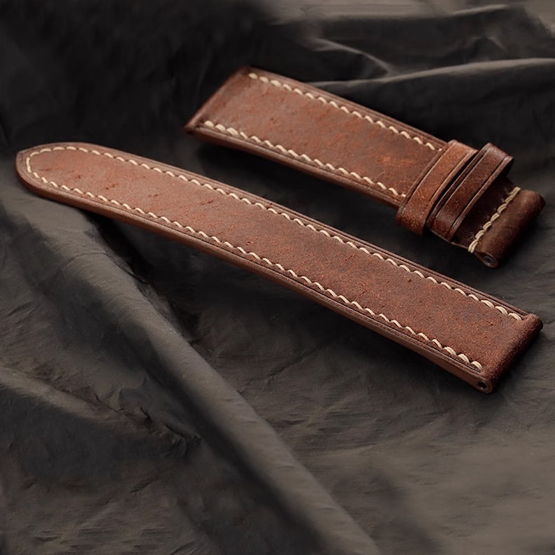 Op de afbeelding: Twee bruine leren horlogebandjes met witte stiksels. De bandjes zijn gemaakt van een distressed leermateriaal en zijn ontworpen om aan een horloge te worden bevestigd. De bandjes worden op een donkere achtergrond getoond.