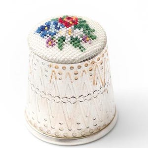 Könnte beinhalten: Ein silberner Fingerhut mit einem Blumendesign auf der Oberseite. Die Oberseite des Fingerhuts ist weiß mit einem Kreuzstichmuster aus blauen, roten, grünen und lila Blumen. Der Körper des Fingerhuts hat ein Zickzackmuster.