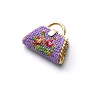 Könnte beinhalten: Eine violette, gestickte Handtaschenbrosche mit Blumenmuster. Die Handtasche ist aus Stoff und hat einen goldenen Metallverschluss.