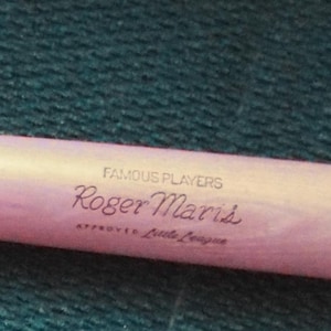 May include: A light pink wooden baseball bat with the Wilson logo and the name "Roger Maris" printed on it. The bat is lying on a green surface.