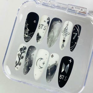 Peut inclure: Un ensemble de dix faux ongles en forme d'amande dans un boîtier en plastique transparent. Les ongles présentent une palette de couleurs noir et blanc avec des accents métalliques argentés, incluant des motifs de pagode, le texte BTS, une lune.